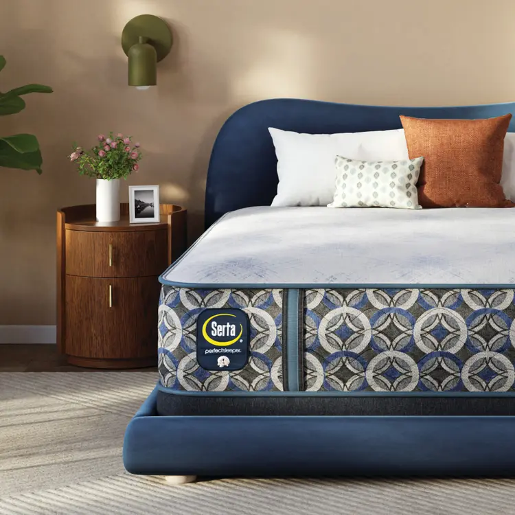 Bed with blue headboard and patterned Serta Perfect Sleeper Hybrid Firm mattress in a bedroom setting.