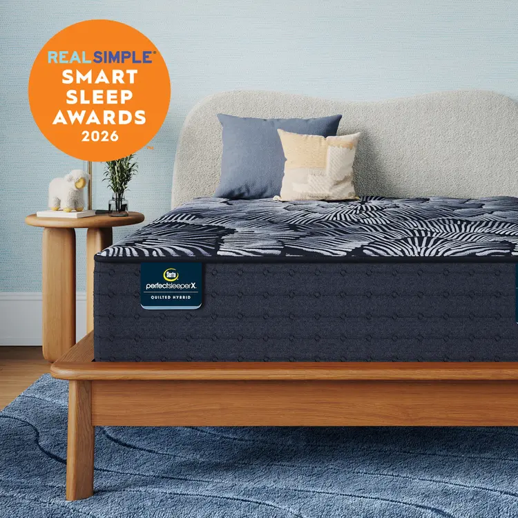 A Perfect Sleeper X Quilted Hybrid Excelled Plush mattress with a dark pattern and matching pillow shams styled in a bedroom setting, with a light wood bed frame, blue rug, side table, and a large orange badge reading 'REAL SIMPLE SMART SLEEP AWARDS 2026'. The mattress label shows 'perfectscore X' and 'BETTER START'.